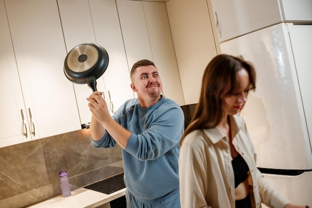 Joking man swinging frying pan toward the head of an unsuspecting woman while they cooking together on modern kitchen at home