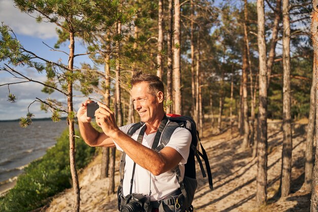 Jolly mature male with backpack is standing in forest near sea shore and using camera on smartphone