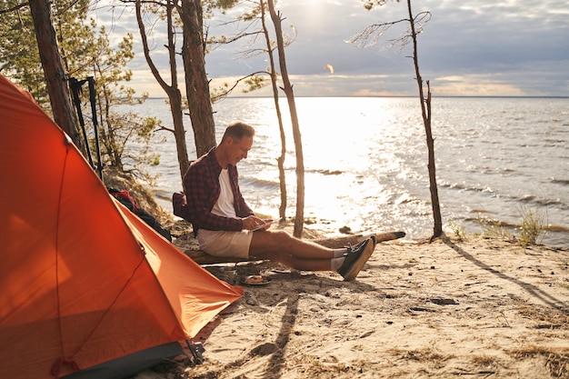 Jolly mature sporty male is sitting near tent in wood next to seaside and surfing internet on touchpad