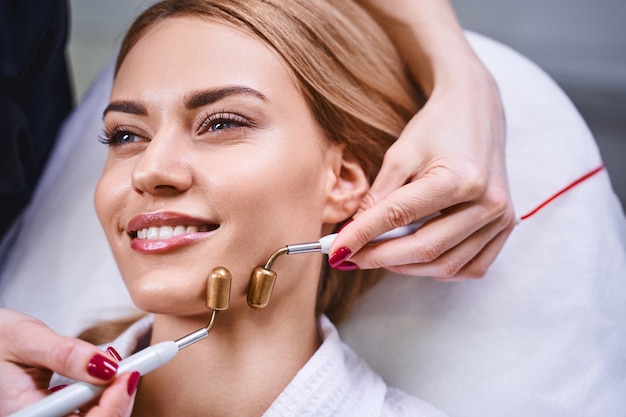 Jolly young woman is relaxing on couch while beauty specialist is using microcurrents treatment on face