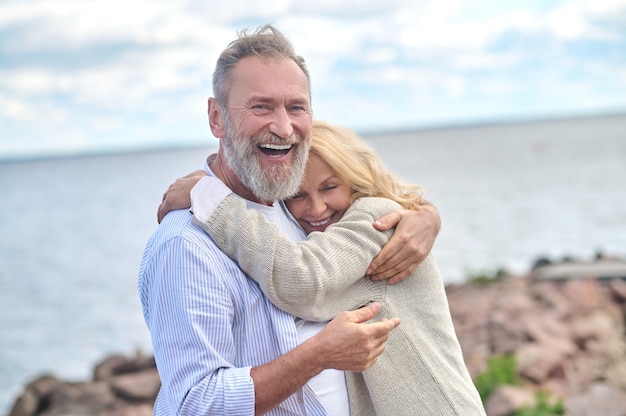 Joy of life. Delighted adult bearded man hugging cute smiling woman standing in nature near sea