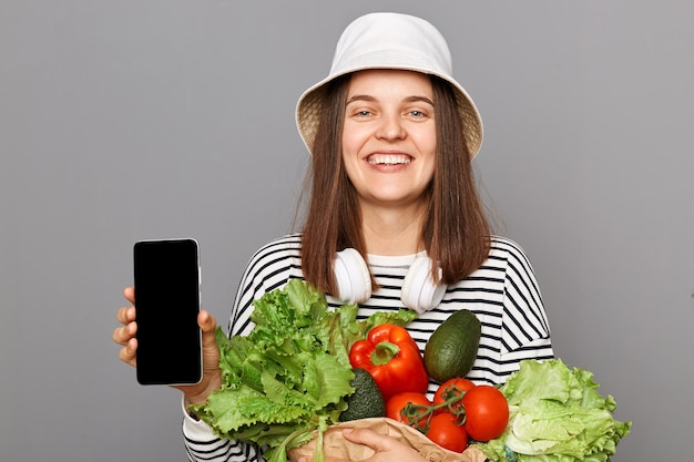 Joyful adorable woman holding vegetables isolated over gray background showing empty screen on smartphone advertisement of organic food delivery