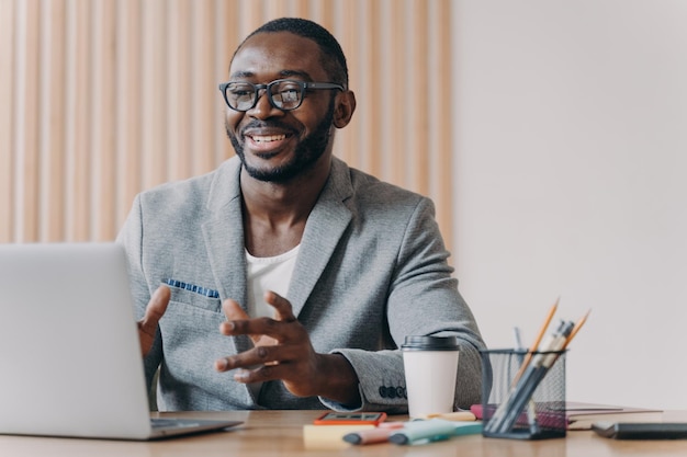 Joyful Afro american businessman wearing glasses and stylish blazer making video call on laptop