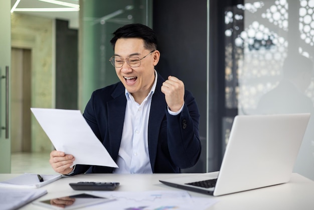 Joyful asian businessman punching the air in success while reviewing documents at his desk with a