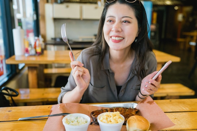 joyful asian female customer holding phone and fork is eating rich breakfast at cozy American restaurant. Holiday morning delightful woman having hearty meal and chatting happily at table.