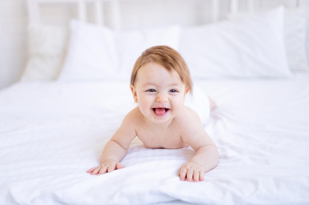 A joyful baby girl of six months on a bed in a diaper crawls and smiles a small child on a cotton bed at home the concept of care and hygiene