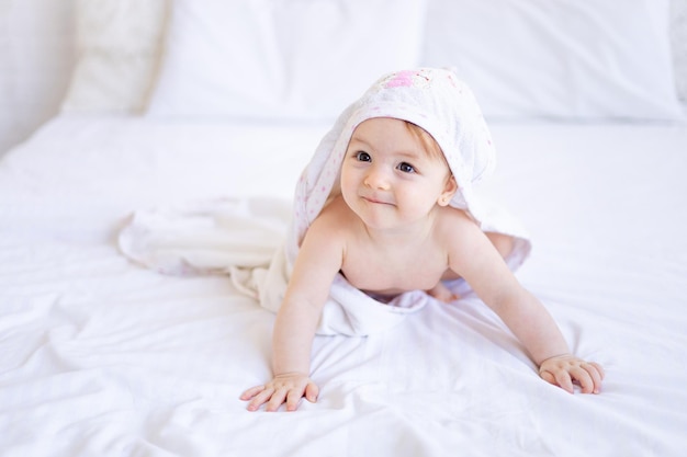 A joyful baby girl of six months on a bed with a white towel on her head after bathing or washing a small child on a cotton bed at home the concept of care and hygiene