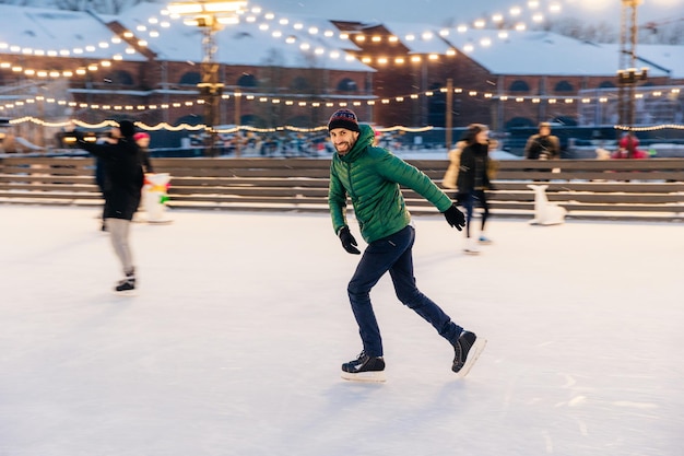 Photo joyful bearded man practices going skating on ice ring has cheerful expression smiles happily demontrates his professionalism active sporty male in green jacket go in for winter sport