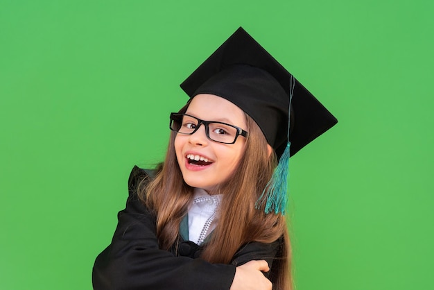 A joyful and beautiful schoolgirl in a graduation cap and a ceremonial robe crossed her arms on an isolated green background. obtaining a certificate in elementary school.