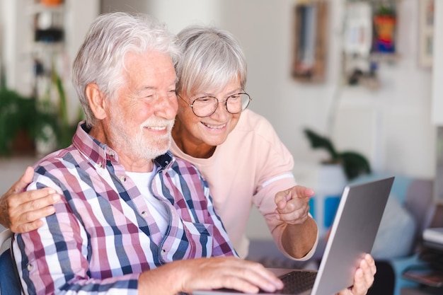 Joyful beautiful senior couple smiling at home browsing together on laptop Active elderly Caucasian couple enjoying technology and social