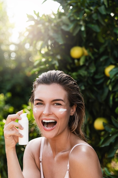 Joyful and beautiful woman applying moisturizing cream or sunblock on her facial skin