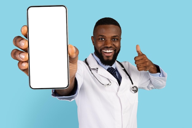 Joyful black doctor demonstrating blank smartphone and showing thumb up at camera