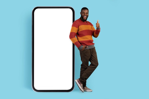 Joyful black man standing near big blank smartphone and showing thumb up