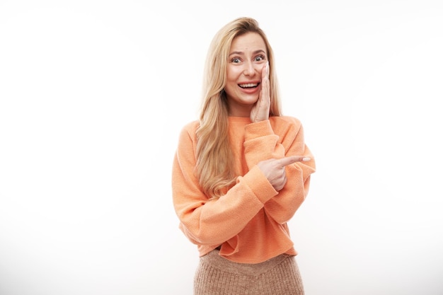 Joyful blonde girl in orange casual smiling points finger at empty copy space for text or product isolated on white background advertising banner