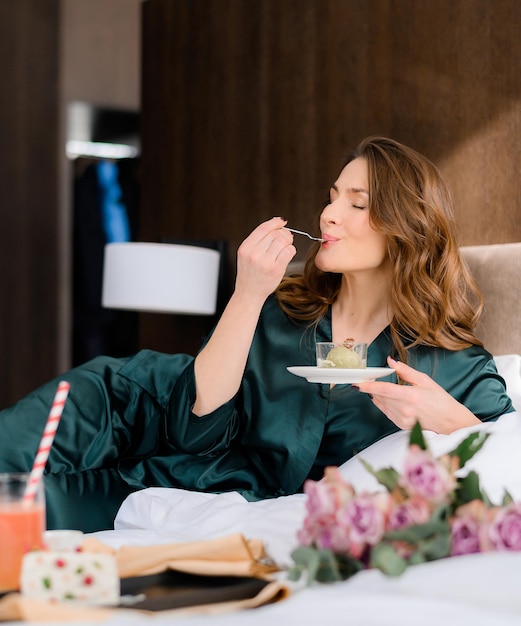 Joyful brunette woman enjoying eating dessert on bed while resting hotel room Attractive lady in stylish pajamas having a day off relaxing alone Luxurious lifestyle Glamorous young woman