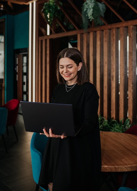 A joyful businesswoman in a black dress works with a laptop in a cafe