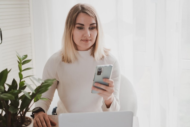 Joyful charming woman using laptop and mobile phone smiling while sitting at the workplace