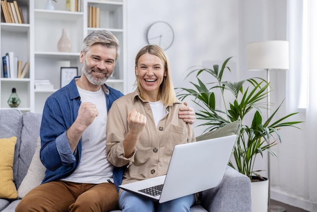 Joyful couple cheering while using a laptop seated on sofa in cozy modern living room expressing