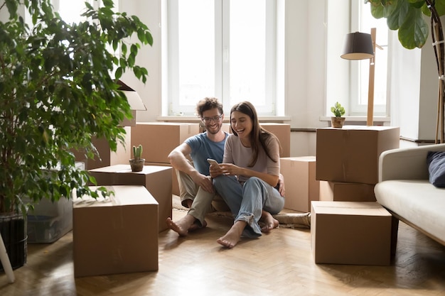 Joyful dating couple moving in apartment together enjoying relocation sitting on floor hugging at heap of paper boxes using online app service on mobile phone smiling laughing