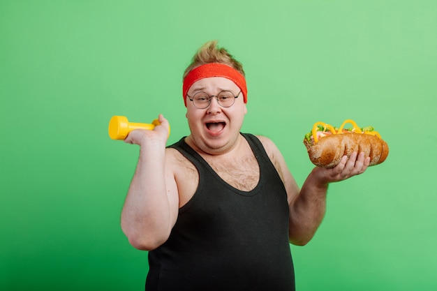 Joyful fat male having fun with burger and dumbbell