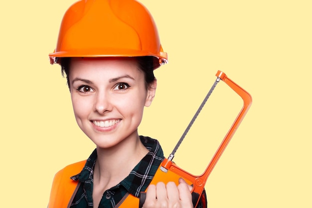 joyful female builder in an orange helmet with a metal saw in her hands yellow background