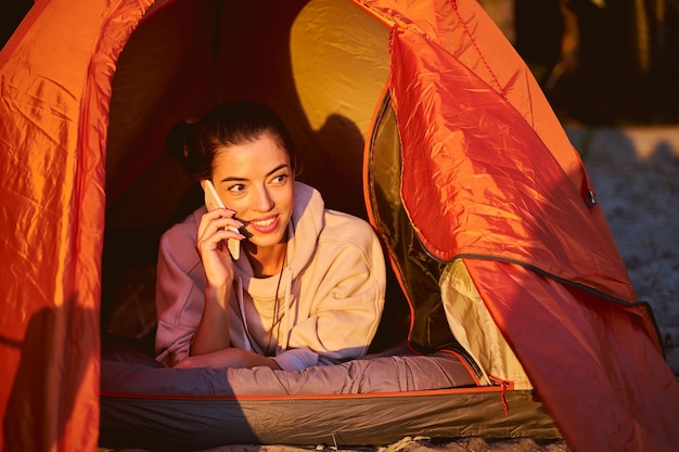 Joyful female traveler having phone conversation and smiling while resting in touristic tent