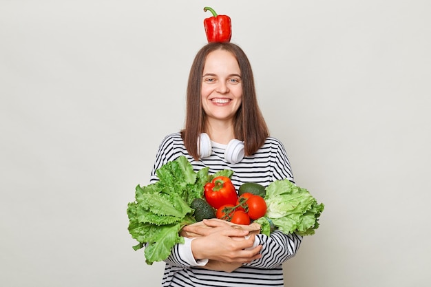 Joyful funny woman with brown hair wearing striped shirt standing isolated over gray background holding vegetables posing with pepper on head looking at camera