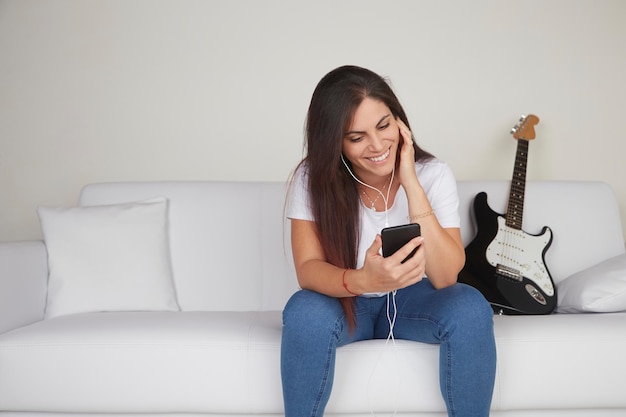 Joyful girl in earphones watching electric guitar tutorial on mobile phone