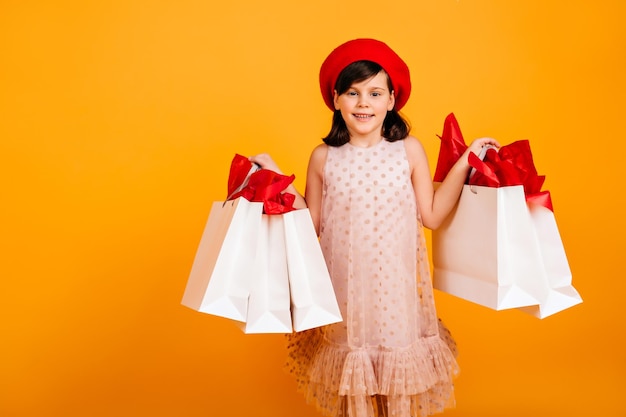 Joyful girl in elegant dress smiling on yellow background Cute child holding store bags and looking at camera