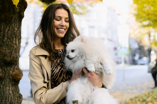 Joyful girl holds a white dog in her arms A young woman holds her dog under a tree with yellow leaves city background