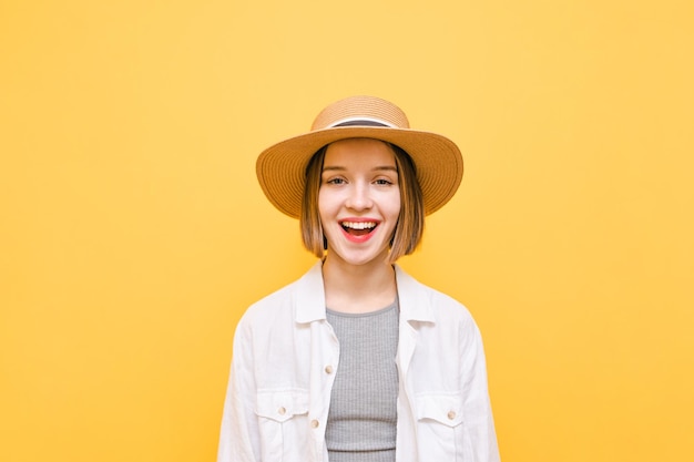 joyful girl in straw hat and white shirt on yellow background looks into camera and smiles