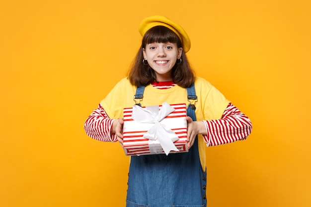 Joyful girl teenager in french beret, denim sundress holding red striped present box with gift ribbon isolated on yellow wall background. People sincere emotions, lifestyle birthday holiday concept.