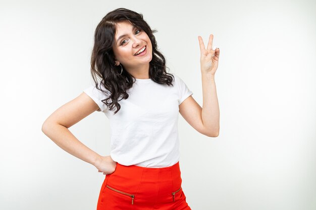 Joyful girl in a white T-shirt shows two fingers on a white background.