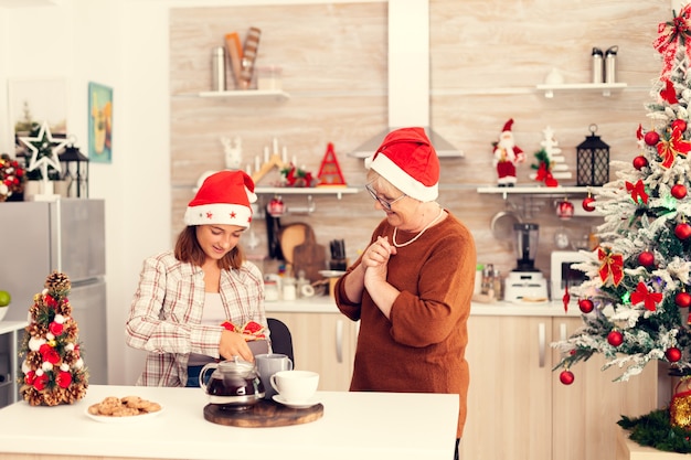 Joyful granny wearing red hat giving niece christmas gift box