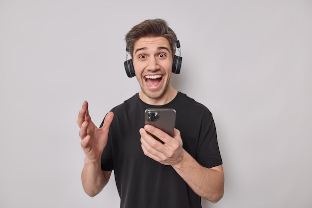 Joyful handsome European guy reacts on amazing news receieves message on his social network page holds mobile phone listens music via headphones isolated over white background feels carefree