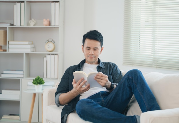 Joyful happy asian man smiling and reading book while sitting on couch sofa in living room at home