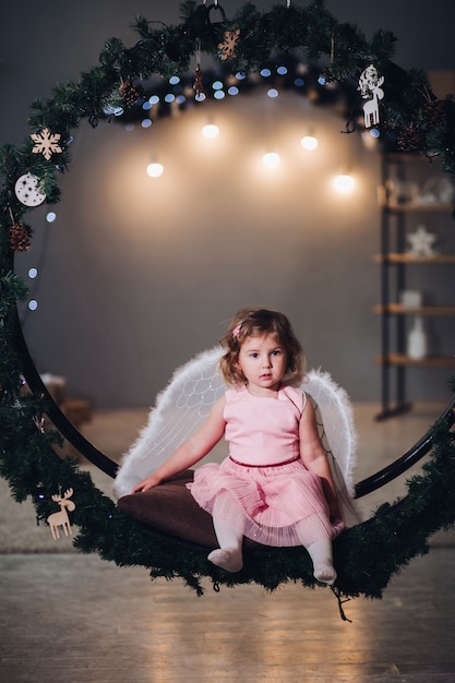 Joyful and happy child sitting and posing on fir wreath decorated with light garland. Adorable girl wearing in cute rose dress and having angel wings behind. Kid smiling and looking at camera.