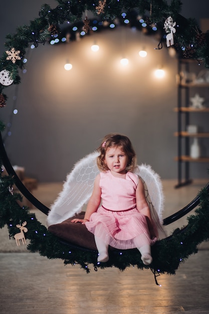 Joyful and happy child sitting and posing on fir wreath decorated with light garland. Adorable girl wearing in cute rose dress and having angel wings behind. Kid smiling and looking at camera.