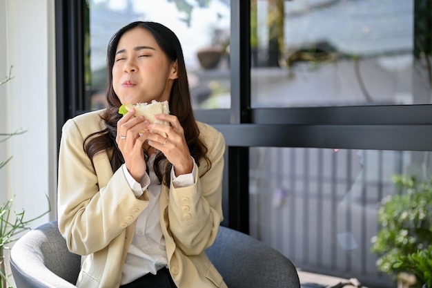 Joyful happy and hungry Asian businesswoman enjoying with her yummy sandwich at the cafe