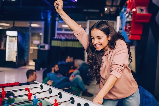 Joyful happy young woman stand in room and play table soccer. She wins. Model hold hand up. She plays alone.