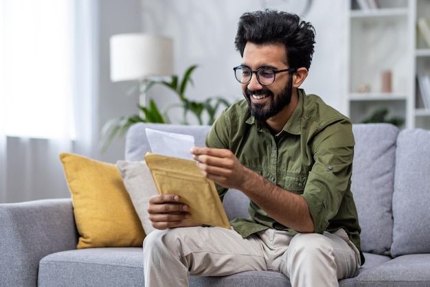 Joyful hispanic received letter mail notification man sitting at home in living room on sofa holding