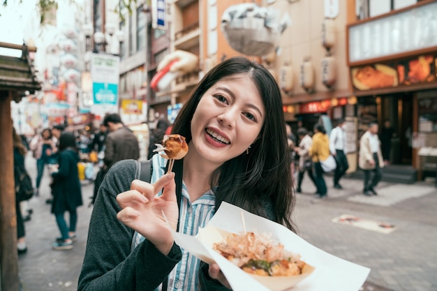 joyful lady traveler showing takoyaki to the camera. young girl cheerful trying japanese street food. travel in dotonbori in osaka japan.