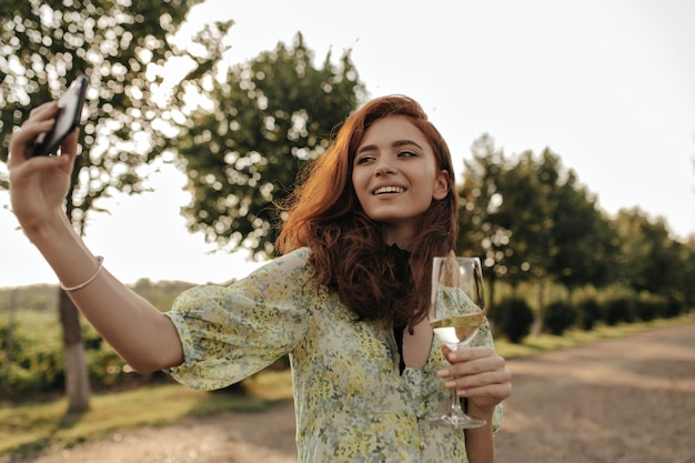 Joyful lady with red hairstyle and bandage on neck in fashionable printed dress making selfie and holding glass with wine outdoor