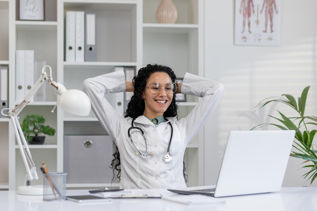 Joyful latina doctor in white coat taking a break at her office desk with laptop feeling content and