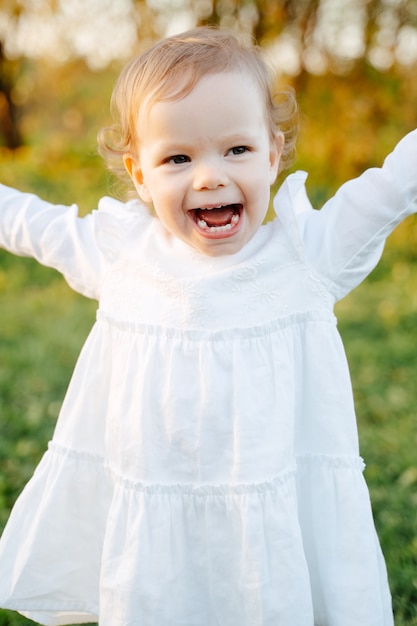 Joyful little girl child in a white cotton dress runs along a green meadow in the field