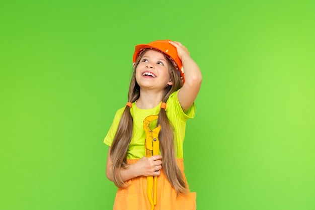 A joyful little girl with an adjustable wrench in her hands looks up and holds a protective helmet with her hand. Repair and construction. Isolated background.