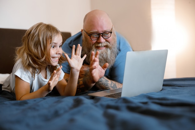 Joyful little girl with funny bearded daddy wave hands to laptop camera at videochat on