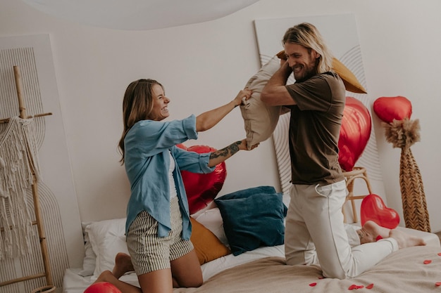 Joyful loving couple having a pillow fight in bed surrounded with red heart shape balloons