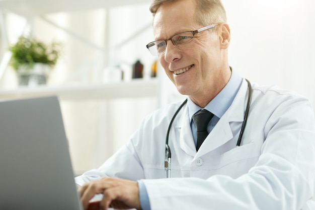 Joyful male doctor working on laptop in clinic