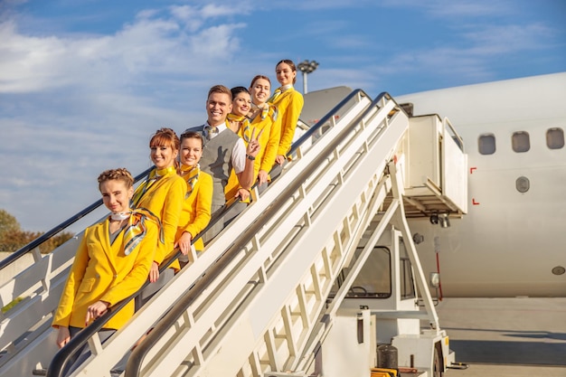 Joyful man and female flight attendants in aviation uniforms looking at camera and smiling while standing on plane boarding stairs